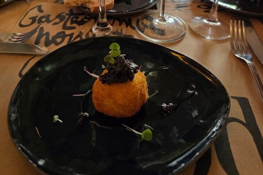 Un plato negro con un alimento esférico cubierto con una sustancia oscura y un adorno verde, sobre una mesa con un papel con texto. 3