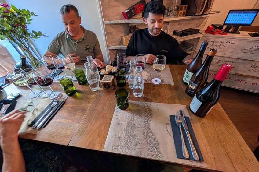 Due uomini seduti a un tavolo di legno, con bicchieri da vino, bottiglie, una mappa e un portatile. 3