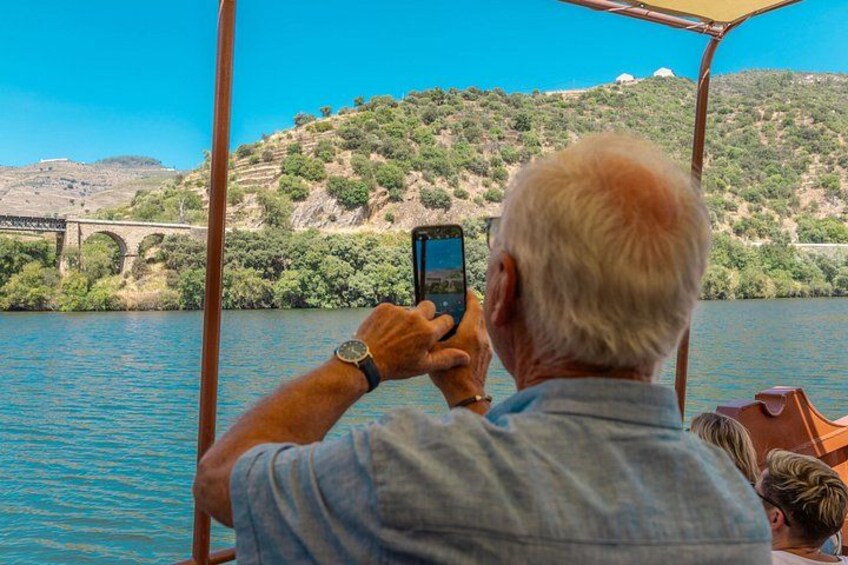 A person taking a photo with a smartphone, a boat, and a bridge in the background. 3