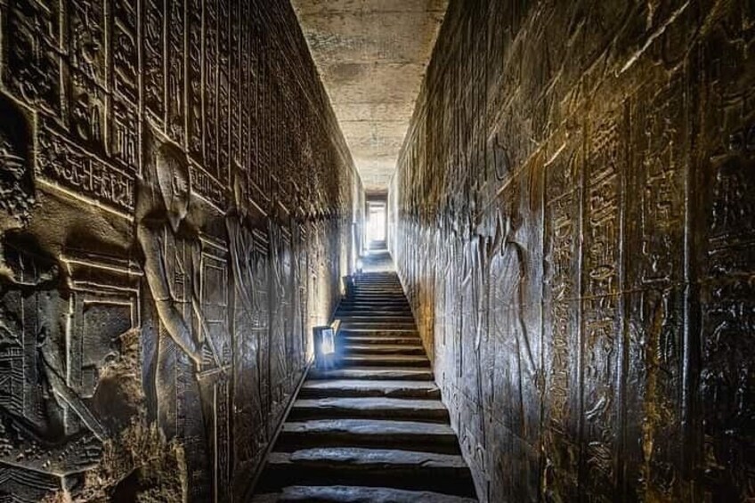 A narrow passageway with hieroglyphs on the walls and a staircase leading to a light source at the end. 4