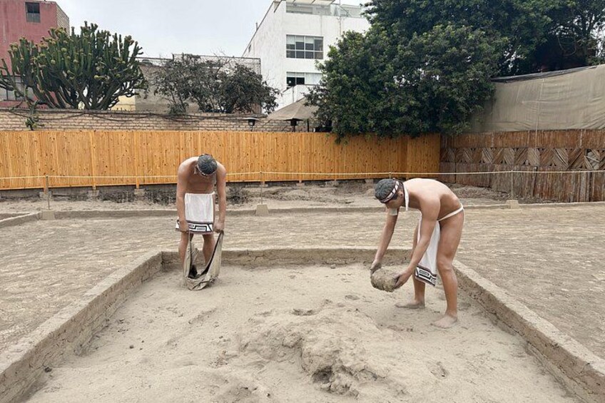 Two people digging in a sandy area with a wooden fence and buildings in the background. 3