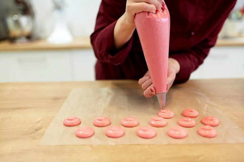 A person using a pink piping bag to decorate macarons on parchment paper. 1