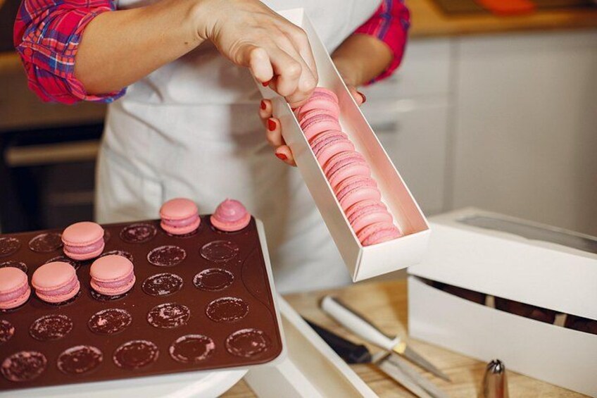 A person in a kitchen, holding a box of pink macarons. 4