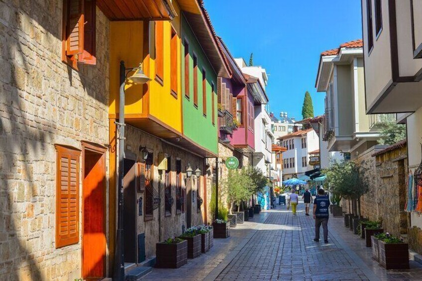 Une ruelle étroite pavée, bordée de bâtiments aux couleurs vives, où l’on aperçoit une personne qui se promène et des plantes en pot le long du trottoir. 5