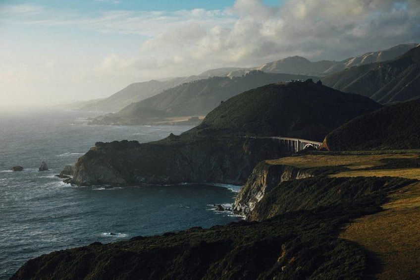 A coastal landscape with a bridge spanning a deep canyon, surrounded by hills and the ocean. 2