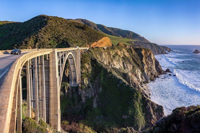 A bridge over a canyon with a road, ocean waves, and green hills. 1