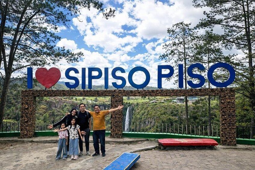 A family posing in front of a sign that reads 'I ♥ SIPISO PISO' with a natural landscape in the background. 3