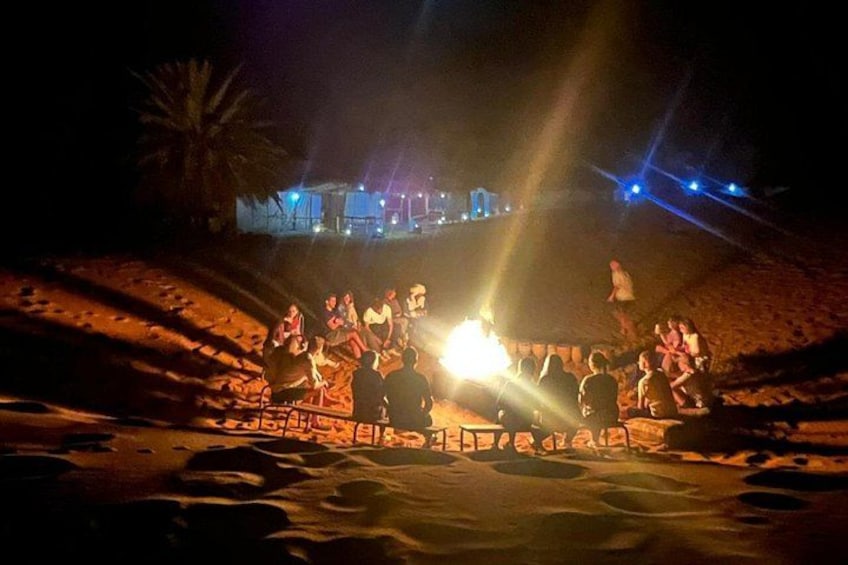 A group of people gathered around a fire at night on a sandy beach with palm trees and a building in the background. 4