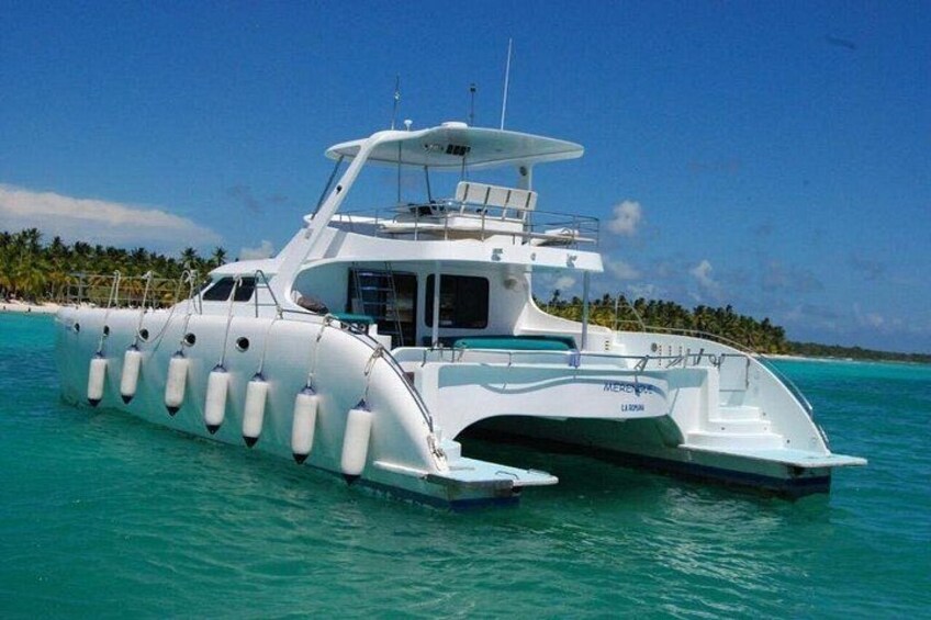 Una embarcación blanca con cabina y cubierta techada, flotando sobre aguas turquesas con una playa y palmeras al fondo. 2