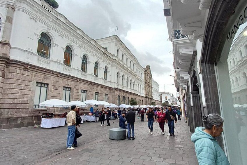 Une rue piétonne bordée de bâtiments blancs, où l’on voit des gens se promener et des étals de marché en plein air. 4