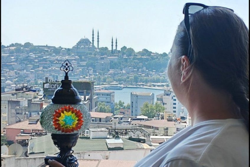 Eine Stadtansicht mit einer markanten Moschee und Minaretten im Hintergrund, einer Zierlampe im Vordergrund und einer Person mit Sonnenbrille, die die Aussicht betrachtet. 3