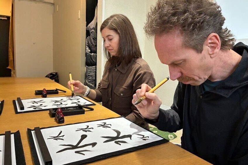 Dos personas practicando caligrafía en una mesa de madera con papel y herramientas de caligrafía. 1