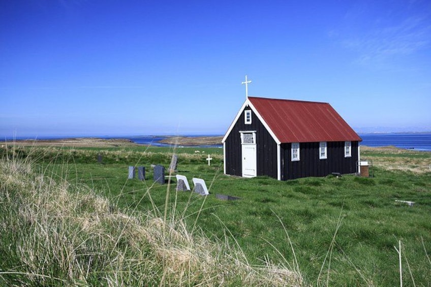 A small black church with a red roof, surrounded by grass and a few gravestones in a coastal landscape. 3