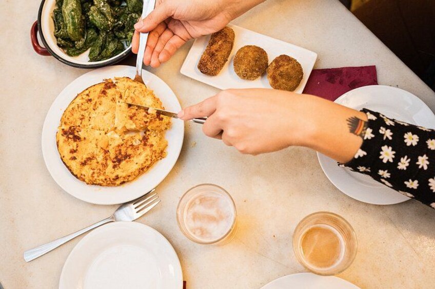 A person cutting a large pancake with a knife on a white plate, with a side dish of rolled vegetables and a plate of fried items. 2