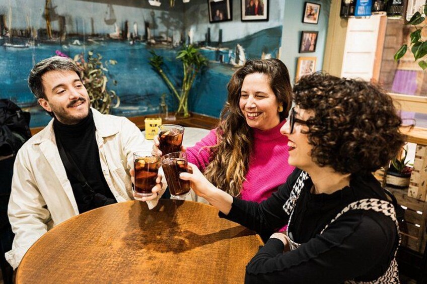 Three people toasting with drinks at a round wooden table in a bar with a blue wall and plants. 4