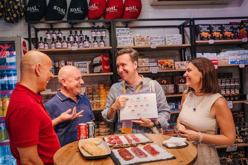 Un groupe de personnes se trouve dans un magasin où les étagères sont remplies de produits, un comptoir présente des articles alimentaires, et un réfrigérateur est visible à l’arrière-plan. 3