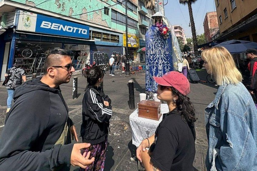 Escena callejera con una persona vestida con un atuendo azul estampado y un gorro rojo en el centro, una tienda 'BENOTTO' a la izquierda, y otra persona con una chaqueta negra y un gorro rosa a la derecha. 4