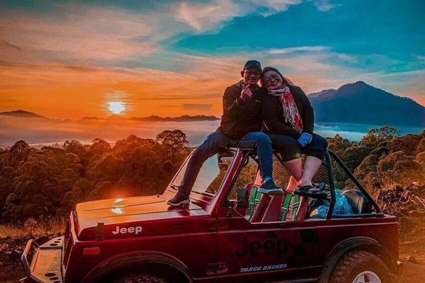Un Jeep rojo con dos personas sentadas en el capó, un atardecer al fondo y un paisaje montañoso. 5