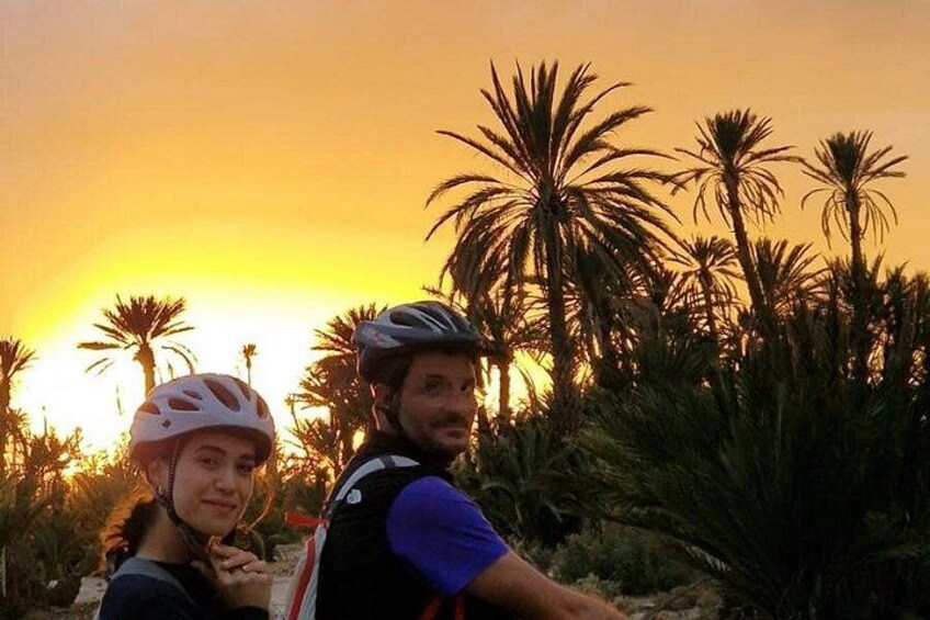 Two people with helmets, one in a white helmet and the other in a black helmet, standing in a natural setting with palm trees and a sunset in the background. 1