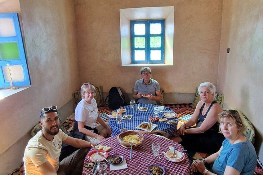 A group of people sitting around a table with food and drinks, in a room with a window and a blue and white checkered tablecloth. 2