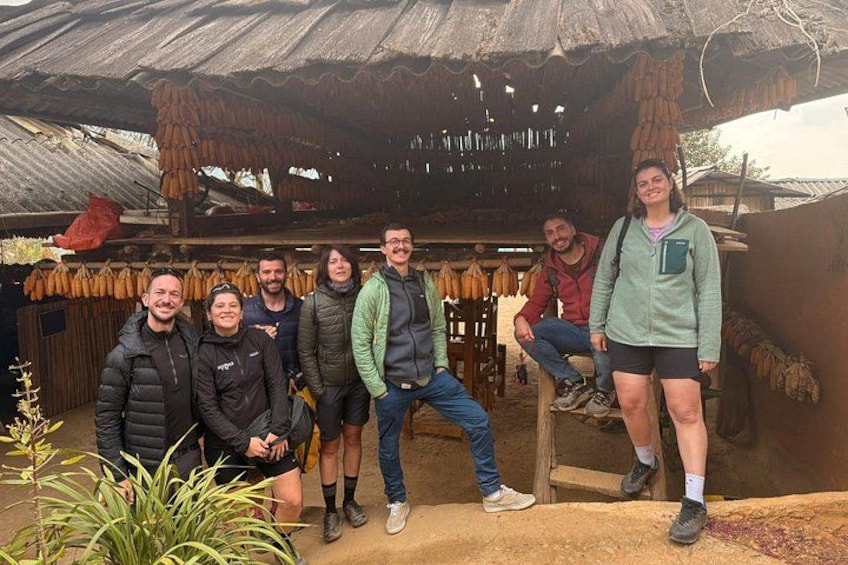 Un groupe de personnes se trouve dans un espace extérieur, devant une structure rustique à toit de chaume, un banc en bois et des produits séchés suspendus. 1
