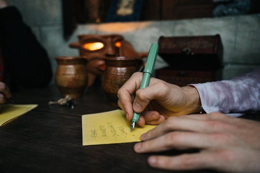 Una persona escribiendo en una nota amarilla con un bolígrafo verde, sobre un fondo rústico que incluye cerámica y una vela. 5