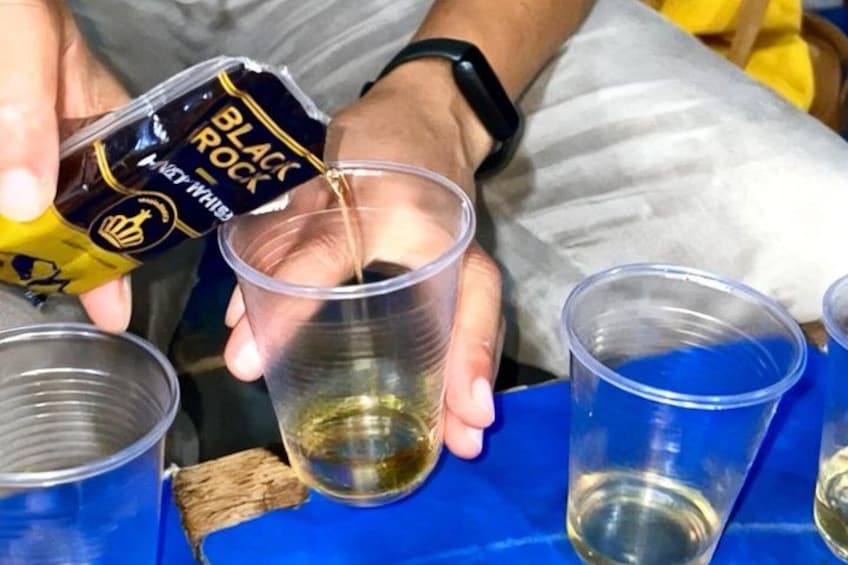 Una persona versa un liquido da una bottiglia etichettata 'BLACK ROCK' in un bicchiere di plastica trasparente. 4