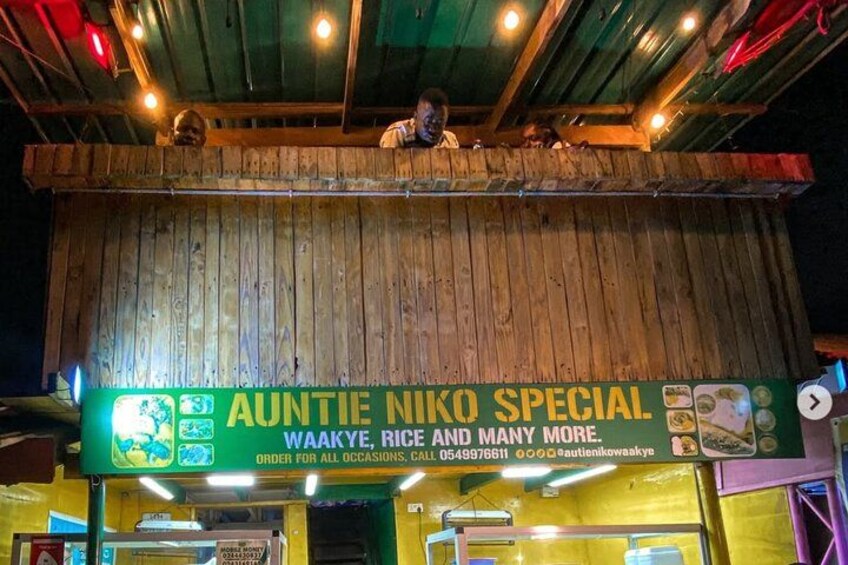 Un restaurant à l’extérieur en bois, avec une enseigne qui fait la publicité de plats spéciaux, notamment « le plat spécial de tante Niko » et « Wakaye, riz et bien d’autres ». 1