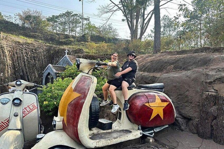 Two people on a vintage scooter with a red and yellow star emblem, parked on a dirt path with a small building in the background. 3