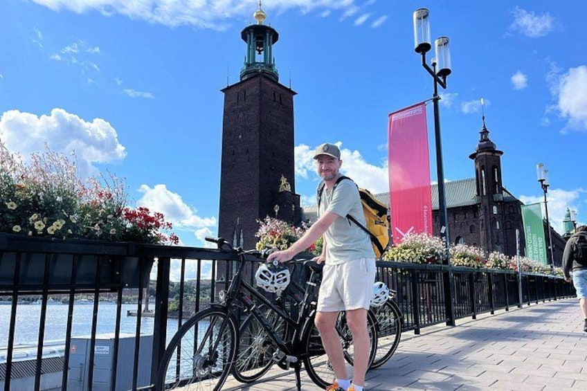 A person standing next to a bicycle with a helmet on the ground, a river, a brick tower, and a red banner in the background. 3