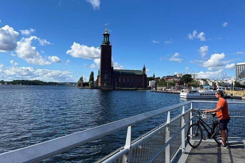 Una persona en un muelle con una bicicleta, una extensa masa de agua, una torre alta y un barco al fondo. 4