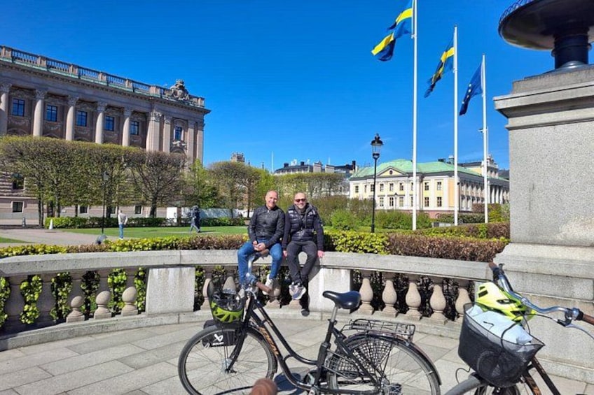 Two people sitting on a bench with bicycles in front of a building with flags. 4