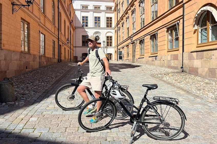 A person standing next to a bicycle on a cobblestone street with buildings in the background. 1