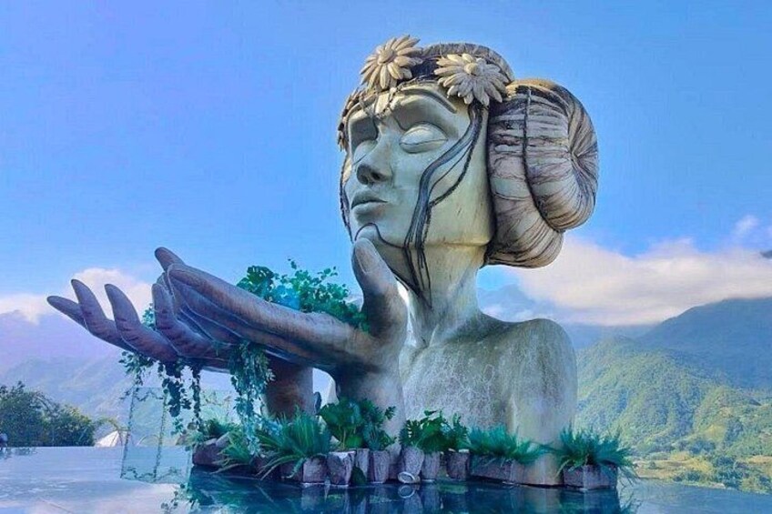 A statue of a woman with a flower crown, hands outstretched, surrounded by water and plants. 1