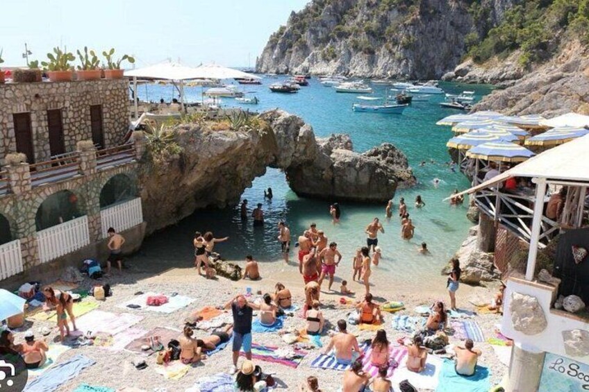 Una playa costera con gente tomando el sol, un arco rocoso y barcos en el agua. 1