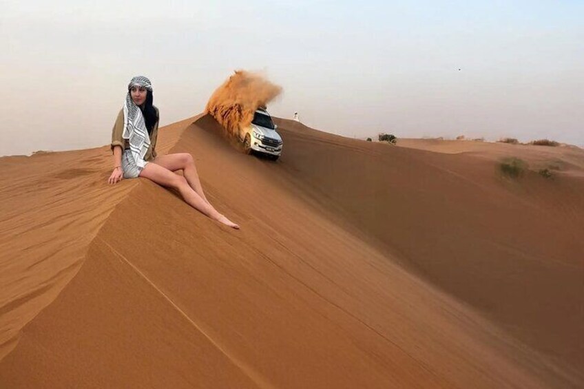 A person sitting on a sand dune with a car driving off-road in the background. 1