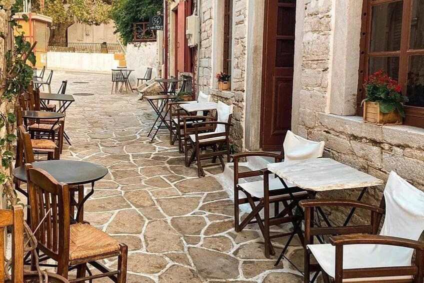 A stone-paved outdoor seating area with wooden tables and chairs, surrounded by stone buildings with windows and doors. 4