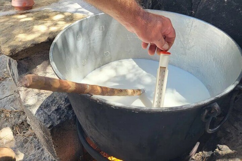 Een hand houdt een thermometer vast in een grote metalen pot gevuld met een witte vloeistof, buitenstaand op een stenen oppervlak. 1