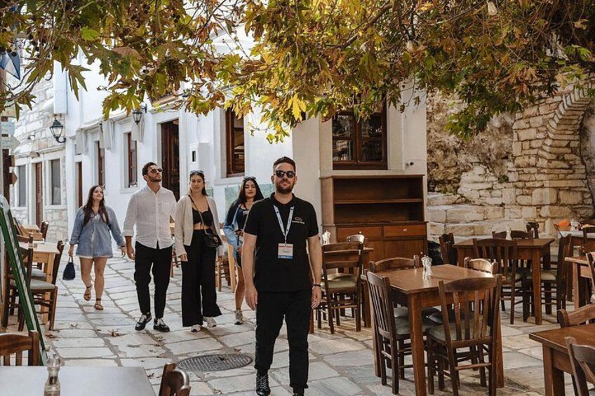 A group of people walking on a cobblestone street with outdoor seating, surrounded by white buildings and stone walls. 5