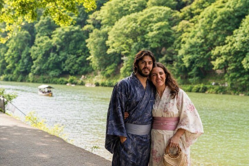 A couple in traditional Japanese attire by a river with lush greenery and a boat in the background. 1