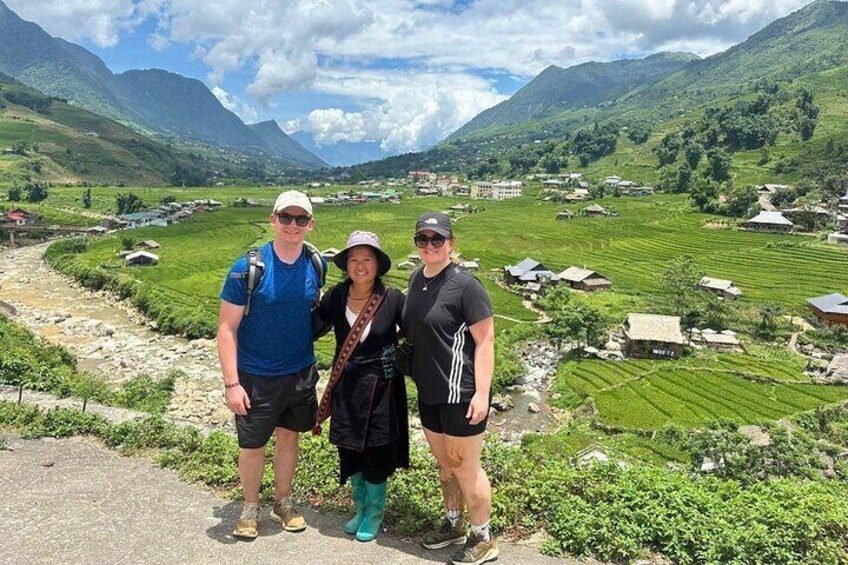 Three people standing on a path with a mountainous landscape and terraced fields in the background. 3