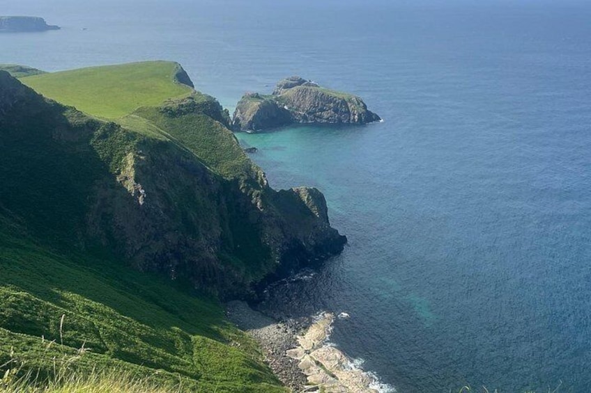 A coastal landscape with green hills, rocky outcrops, and clear blue water. 1