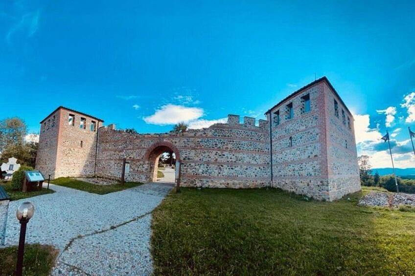 A historic stone building with a central archway, surrounded by a gravel path and grassy area. 1