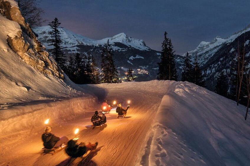 A snowmobile trail with riders, snow-covered mountains, and a lit-up town in the background. 1