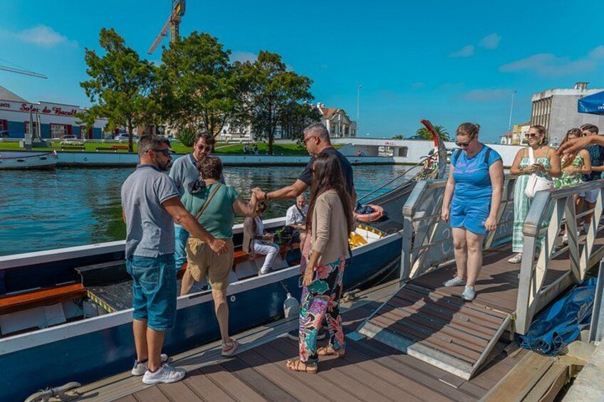 Un gruppo di persone su un molo di legno, vicino a un corso d'acqua, con barche e un edificio sullo sfondo. 3