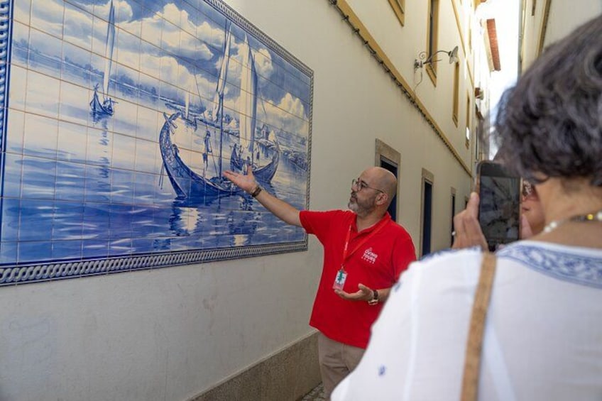 A tiled wall with a maritime scene, a person in a red shirt gesturing towards it, and another person taking a photo. 5