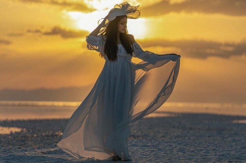 Eine Person in einem weißen, fließenden Kleid steht an einem Strand, im Hintergrund ein Sonnenuntergang. 5