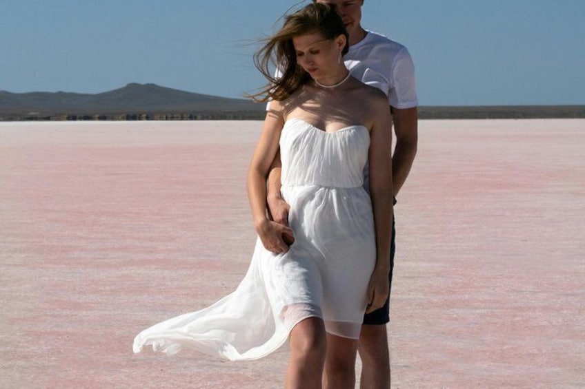 Eine Person in einem weißen Kleid steht auf einer rosa Salzwüste, im Hintergrund eine Berglandschaft. 2