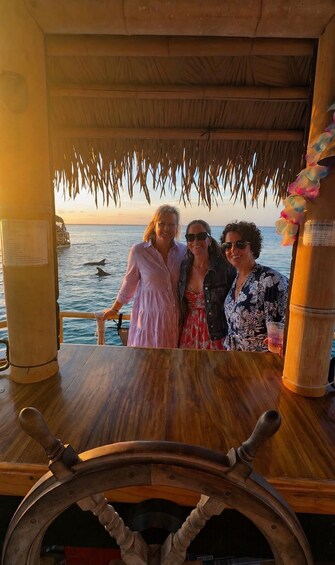 Three people on a boat with a thatched roof, a wooden table, and a steering wheel. 3