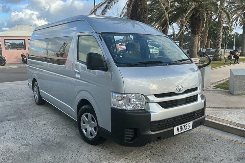 Ein silberner Toyota-Van steht auf einer gepflasterten Fläche, im Hintergrund sind Palmen und ein Gebäude zu sehen. 1